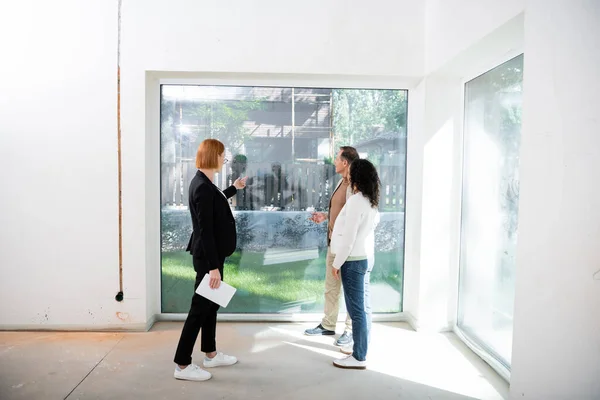 Pelirroja agente inmobiliario en gafas celebración tableta digital, mientras que apunta a la ventana panorámica cerca de pareja interracial dentro de la nueva casa - foto de stock