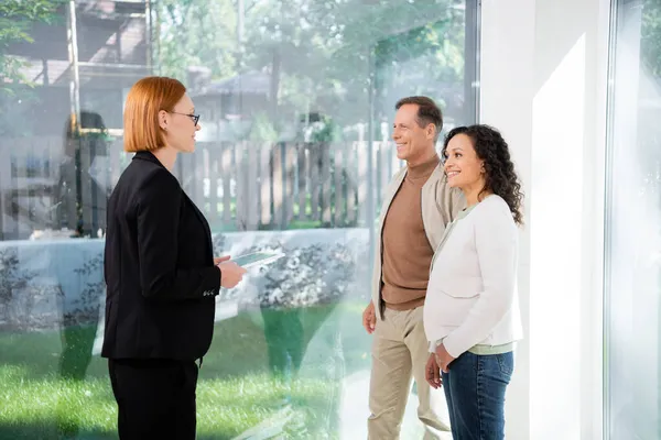 Alegre pelirroja agente inmobiliario en gafas celebración tableta digital cerca de feliz pareja interracial dentro de la nueva casa - foto de stock