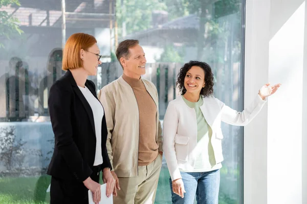 Redhead realtor in glasses standing near african american woman pointing with hand inside of new house — Stock Photo