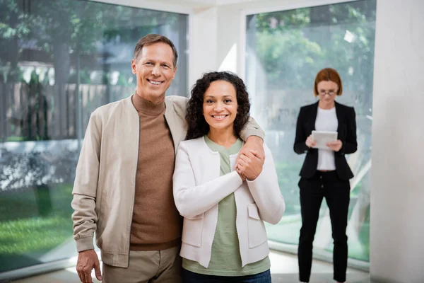 Heureux couple interracial souriant près rousse agent immobilier dans des lunettes tenant tablette numérique sur fond flou — Photo de stock