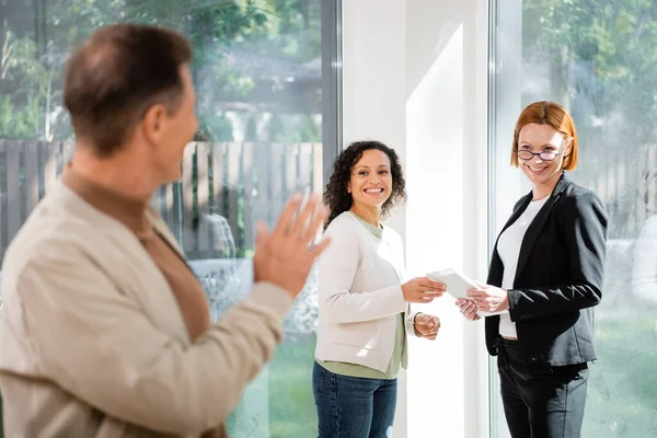 Happy african american woman standing with redhead realtor while looking at blurred husband waving hand in new home — Stock Photo