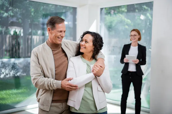 Happy interracial couple hugging near redhead realtor in glasses holding digital tablet on blurred background — Stock Photo