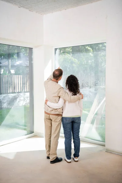 Vista trasera del marido apuntando con el dedo a las ventanas panorámicas mientras abraza a la esposa en una casa nueva - foto de stock