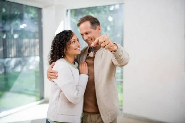 Розмитий і веселий чоловік тримає ключ, обіймаючи афроамериканську дружину в новому будинку — стокове фото