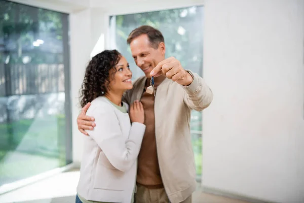 Blurred and cheerful man holding key while hugging african american wife in new house — Stock Photo
