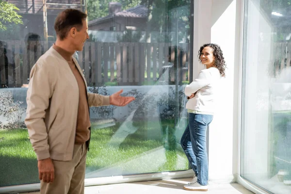Afroamerikanerin steht in der Nähe von Panoramafenstern und sieht verschwommenen Ehemann in neuem Haus an — Stockfoto