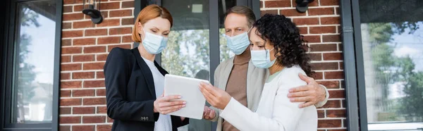Redhead realtor in medical mask showing digital tablet to interracial couple near new house, banner — Stock Photo