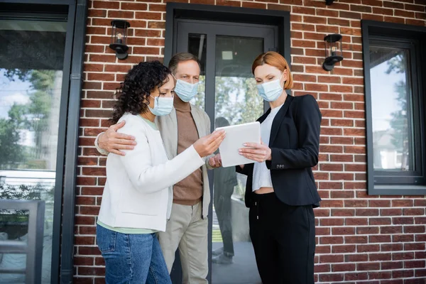 Pelirroja agente inmobiliario en máscara médica mostrando tableta digital a pareja interracial cerca de nueva casa - foto de stock