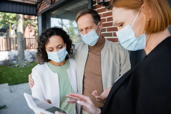 Blurred realtor in medical mask showing digital tablet to interracial couple near new house — Stock Photo