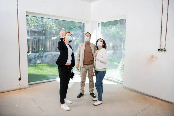 Рудий ріелтор в медичній масці вказує рукою в новому будинку, стоячи біля міжрасової пари — стокове фото