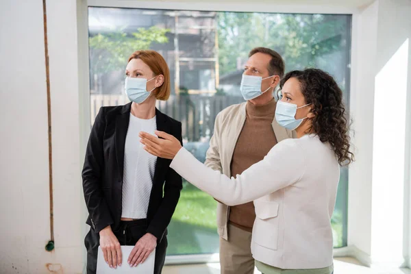 Afrikanerin zeigt mit der Hand in ihr neues Haus in der Nähe von Ehemann und Makler in medizinischen Masken — Stockfoto