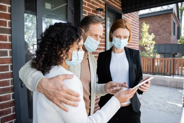 Hombre en máscara médica apuntando a la tableta digital cerca de la esposa afroamericana y agente inmobiliario - foto de stock