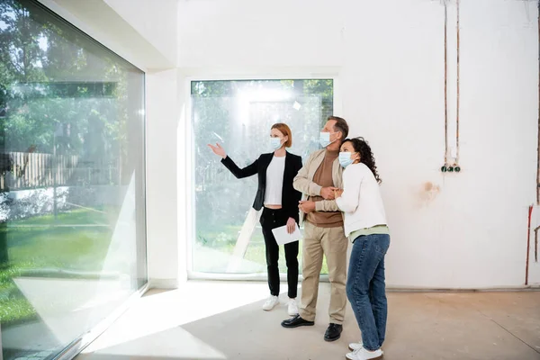 Redhead realtor in medical mask holding digital tablet and pointing at panoramic windows near interracial man and woman — Stock Photo