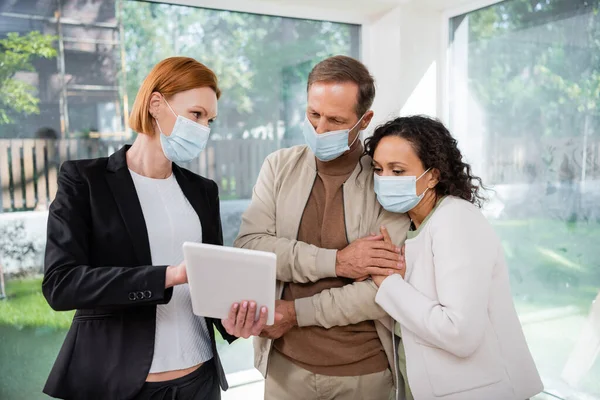 Rothaarige Maklerin in medizinischer Maske zeigt interrassischen Paaren digitales Tablet in neuem Haus — Stockfoto