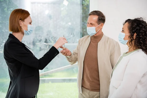 Rotschopf-Makler in medizinischer Maske gibt Schlüssel an exotisches Paar in neuem Haus — Stockfoto