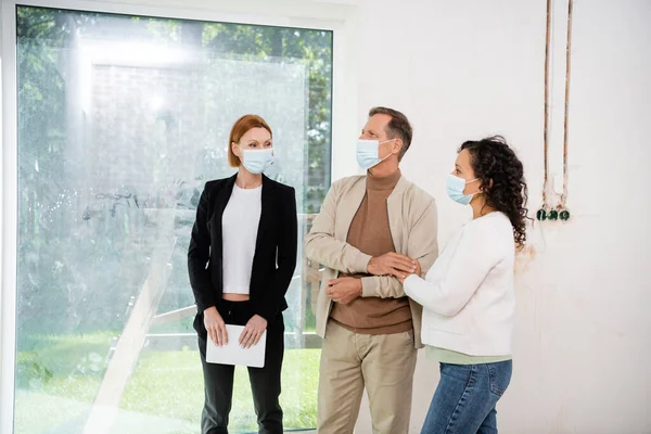 Rousse agent immobilier dans le masque médical tenant tablette numérique et regardant les clients interracial — Photo de stock