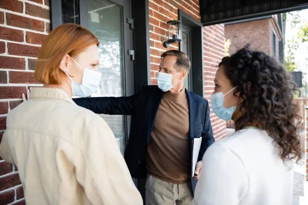 Lesbianas interracial mujeres en médico máscaras mirando uno al otro cerca de bienes raíces y nueva casa - foto de stock