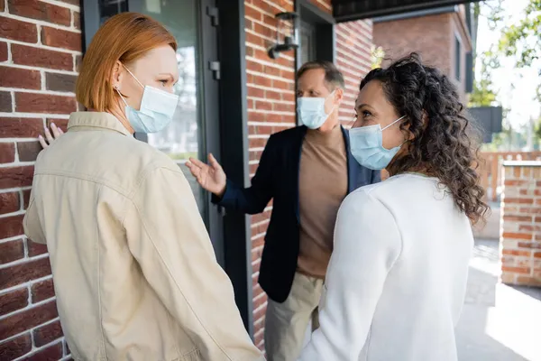 Lesbiche donne interrazziale in maschere mediche guardando l'un l'altro vicino sfocato agente immobiliare e nuova casa — Foto stock