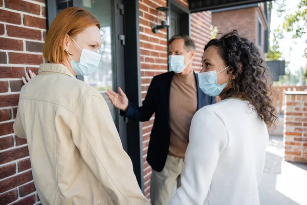 Lesbian interracial women in medical masks looking at each other near blurred realtor and new house — Stock Photo