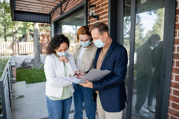 Realtor holding folder with agreement near lesbian interracial couple in medical masks while showing new house — Stock Photo