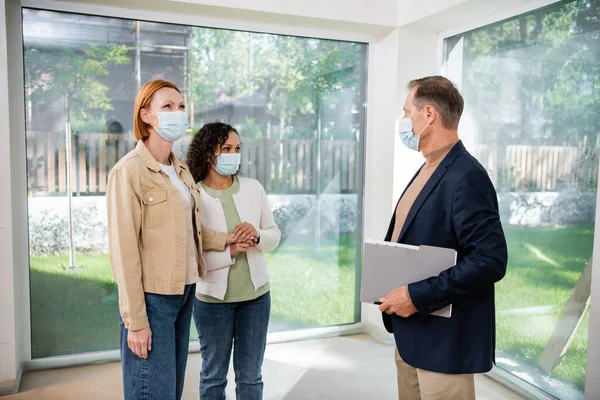 Realtor in medical mask holding folder near interracial lesbian couple inside of new house — Stock Photo