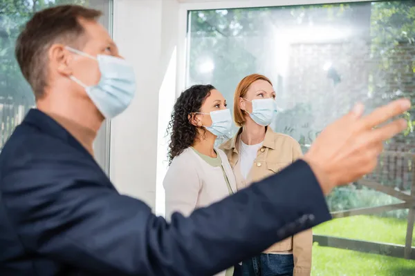 Verschwommener Makler in medizinischer Maske zeigt mit der Hand in die Nähe eines lesbischen Paares in einem neuen Haus — Stockfoto