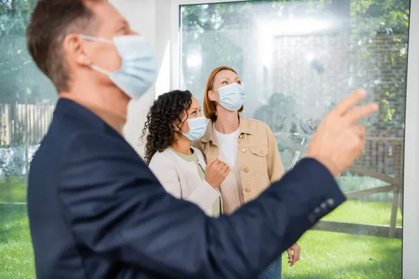 Pareja lesbiana interracial mirando hacia otro lado mientras borrosa agente inmobiliario en máscara médica señalando con la mano en la casa moderna - foto de stock