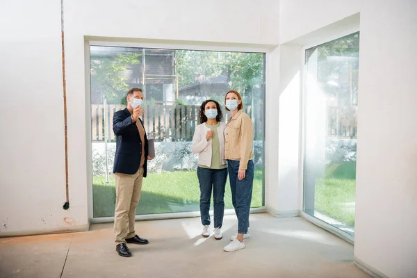 Middle aged realtor in medical mask pointing with hand near interracial lesbian couple inside of new house — Stock Photo