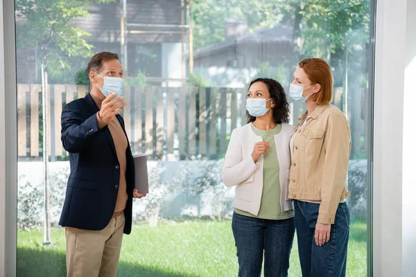 Middle aged realtor pointing with hand near interracial lesbian couple in medical masks inside of new house — Stock Photo