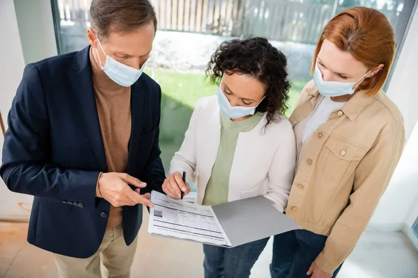 African american woman in medical mask signing contract near realtor and redhead girlfriend — Stock Photo