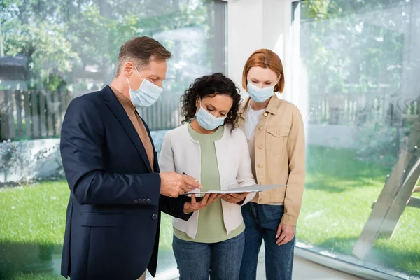 African american woman in medical mask signing contract near middle aged realtor and girlfriend — Stock Photo
