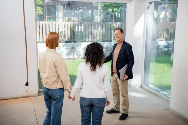 Lächelnder Makler hält Ordner in der Nähe eines lesbischen Paares in einem neuen Haus — Stockfoto