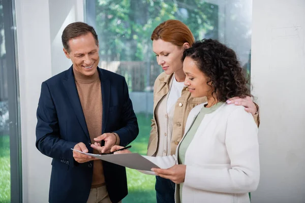 Fröhliche afrikanisch-amerikanische Frau unterzeichnet Vertrag in der Nähe von Makler und rothaarige Freundin — Stockfoto