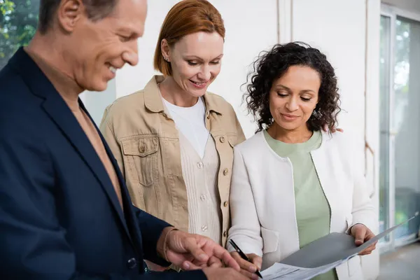 Alegre africana americana mujer firma contrato cerca borrosa agente de bienes raíces y feliz pelirroja novia - foto de stock