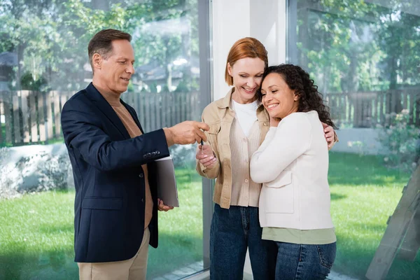 Smiling realtor giving key to interracial lesbian couple inside of new house — Stock Photo