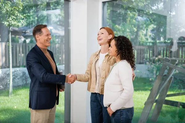 Glücklicher Makler beim Händeschütteln mit lächelnder rothaariger Frau in der Nähe der afrikanisch-amerikanischen Freundin — Stockfoto