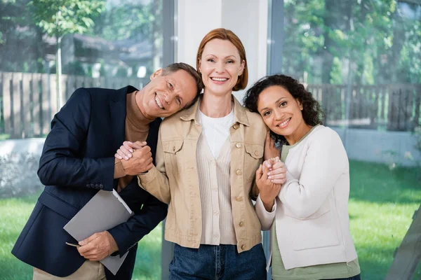 Happy realtor holding hands with cheerful and interracial lesbian couple — Stock Photo