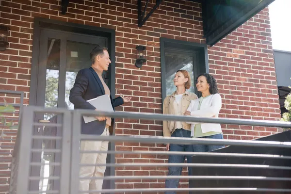 Smiling realtor pointing at house with brick wall near interracial lesbian couple — Stock Photo