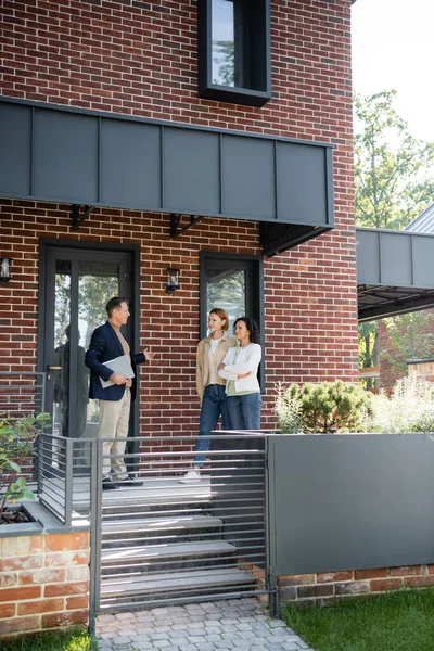 Agent immobilier d'âge moyen pointant vers la maison avec mur de briques près de couple lesbien interracial — Photo de stock