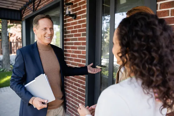 Happy middle aged realtor pointing at house near interracial lesbian couple — Stock Photo