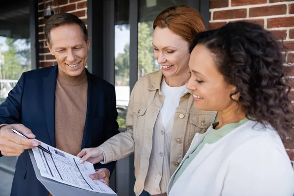 Middle aged realtor holding folder with loan application near happy and interracial lesbian couple — Stock Photo