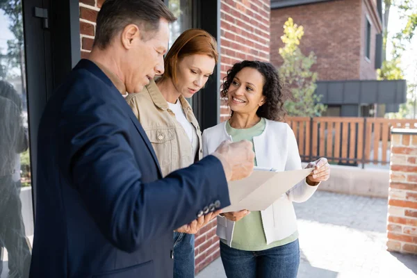 Middle aged realtor holding folder near smiling interracial lesbian couple — Stock Photo