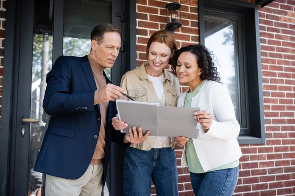 Middle aged realtor holding folder near happy and interracial lesbian couple — Stock Photo