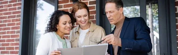 Middle aged realtor holding folder near happy and interracial lesbian couple, banner — Stock Photo