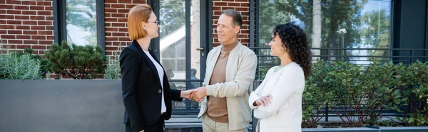 Happy realtor in glasses shaking hands with smiling man standing near african american wife and modern house, banner — Stock Photo