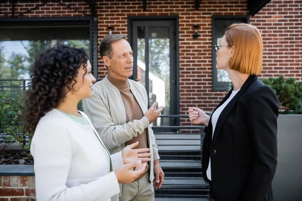 Redhead realtor in glasses talking with multiethnic couple near modern house — Stock Photo