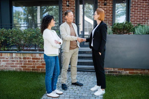 Happy realtor in glasses shaking hands with smiling man standing near african american wife and modern house — Stock Photo