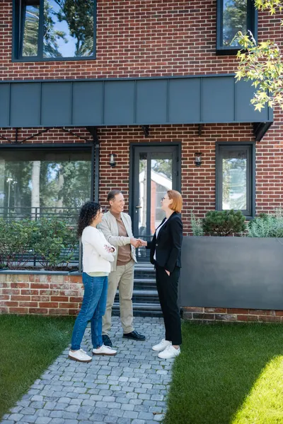Joyeux agent immobilier dans des lunettes serrant la main avec un homme souriant debout près de femme afro-américaine et maison moderne — Photo de stock