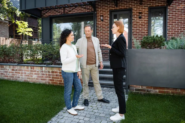 Agent immobilier dans des lunettes geste tout en parlant avec une femme afro-américaine près de mari et maison moderne — Photo de stock