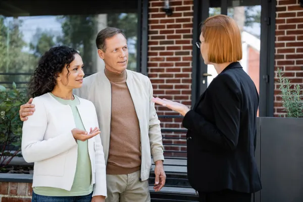 Blurred realtor in glasses gesturing while talking with multiethnic couple near modern house — Stock Photo